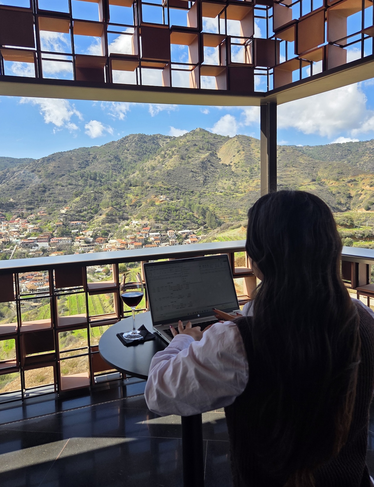 A person with long hair works on a laptop at a table with a glass of red wine, overlooking a scenic view of green hills and a small town through a large open window with a grid-like frame.