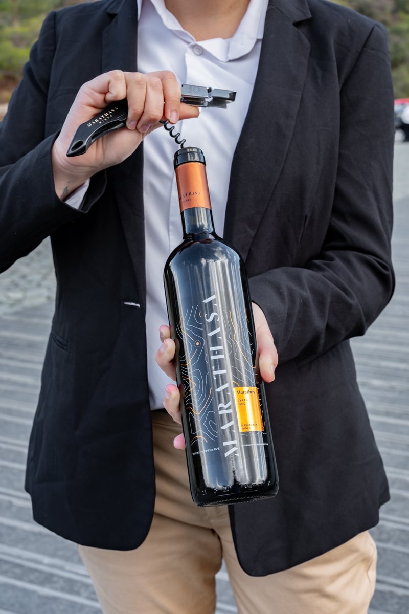 A person in a black blazer and white shirt is holding a bottle of red wine labelled 
