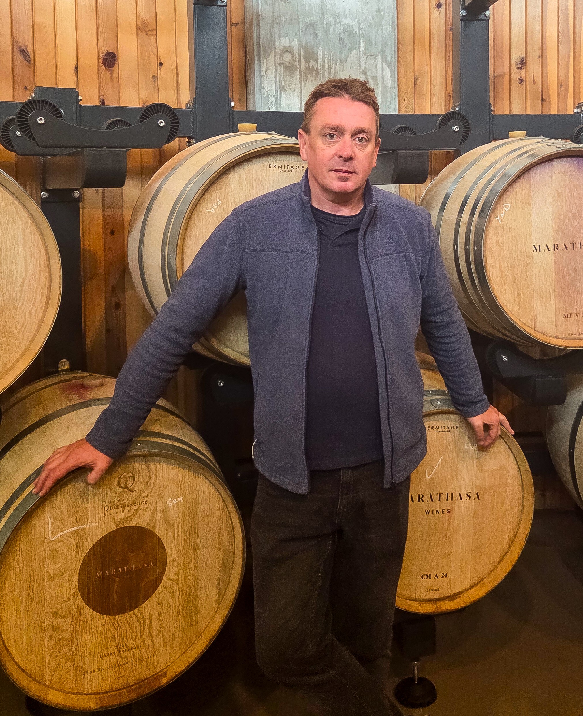A man in a blue jacket stands in front of large wooden wine barrels stacked in a wine vineyard's winery, with one arm resting on a barrel and a wood-panelled wall in the background.