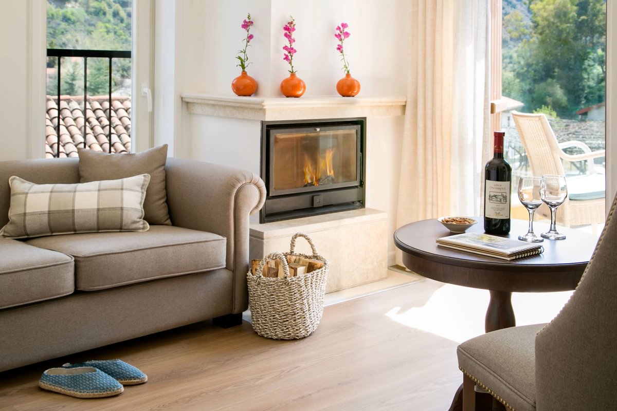 A cosy living room with a beige sofa, fireplace topped with pink flowers, basket of firewood, round table featuring the Culture & Wine set, and sunlight streaming through large windows.