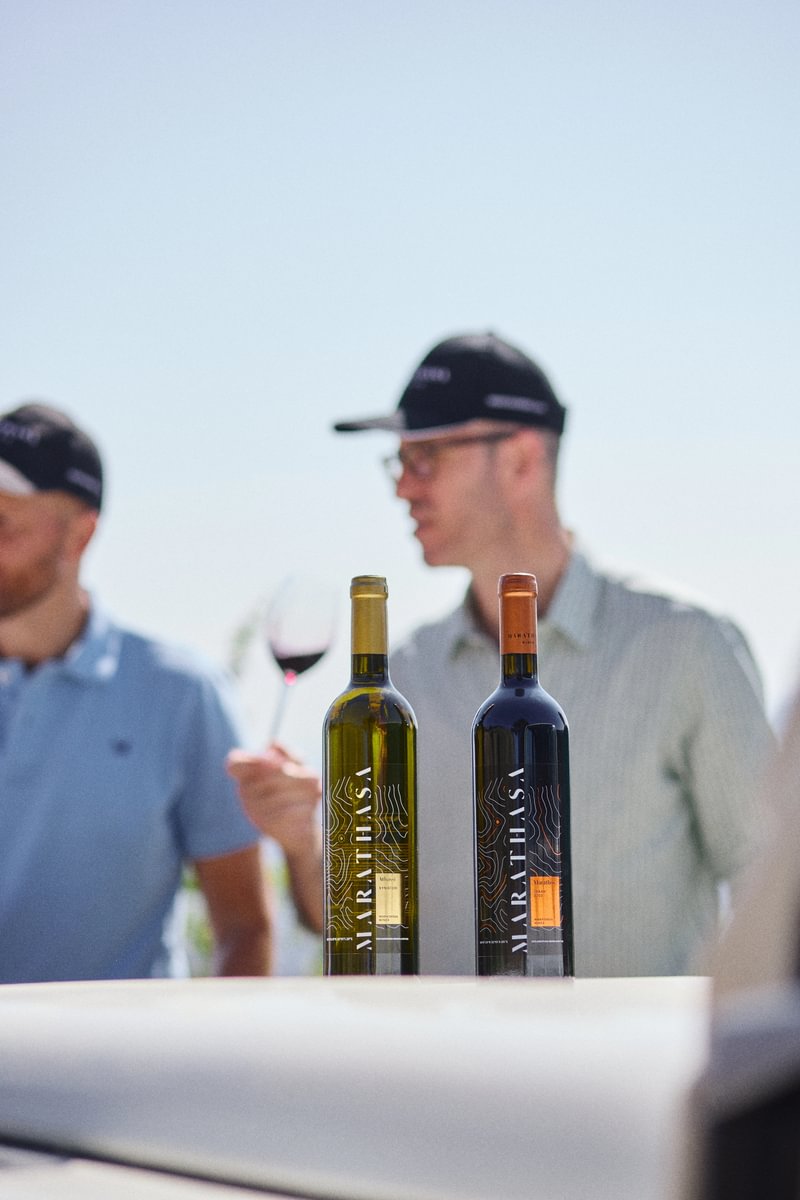 Two bottles of wine are displayed on a white surface outdoors, with two men in casual clothing and black caps in the background, one holding a glass of red wine. The scene is brightly lit with a clear sky.